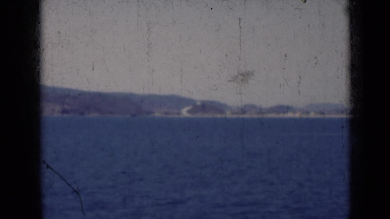 Soft 8mm view of an Indian coastline in 1947, with blue water in front and hazy hills along the horizon.