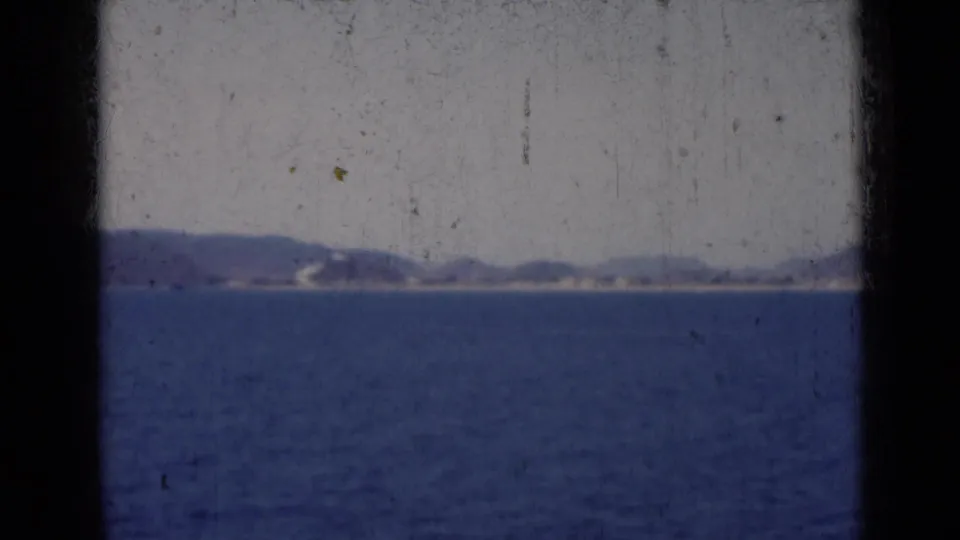 1947 India coastal travel frame with open water, faint sandy shore, and low hills softened by haze and 8mm grain.