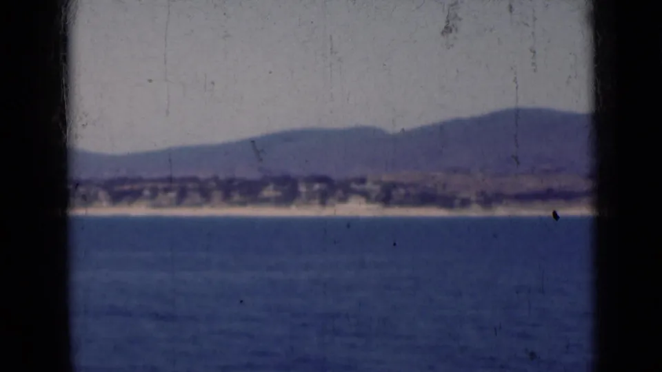 Offshore 1947 India 8mm frame showing blue water, a sandy coast, distant structures, and hazy hills behind them.