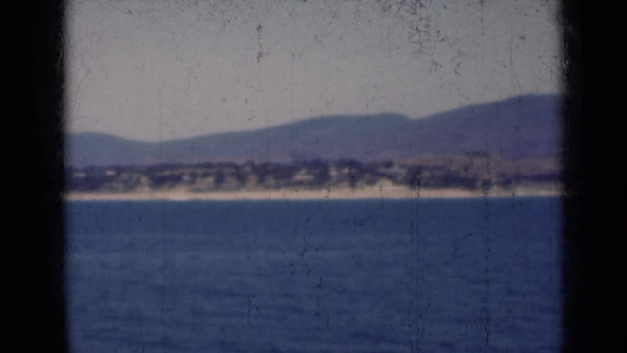 1947 India 8mm frame showing calm water, a distant shore, and hazy mountain ridges viewed through dark vertical foreground obstructions.