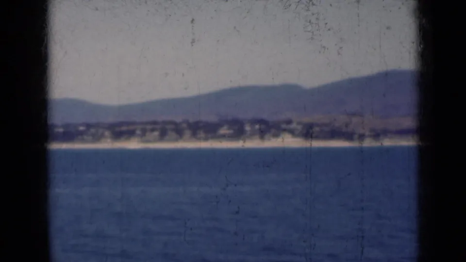 1947 India 8mm frame showing calm water, a distant shore, and hazy mountain ridges viewed through dark vertical foreground obstructions.