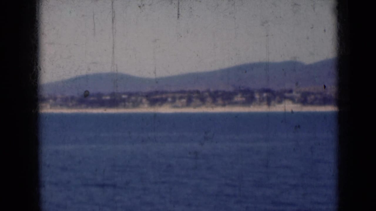 1947 India 8mm frame with calm water in the foreground and a hazy mountain-backed shoreline beyond dark framing bars.