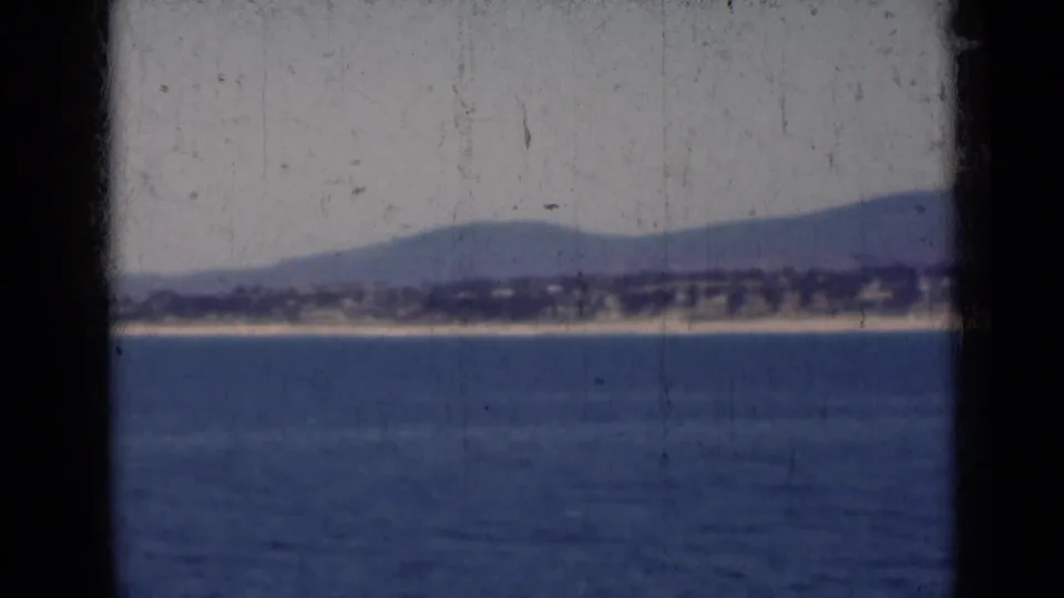 1947 India 8mm frame showing a quiet lakeside or coastal view with blue water, distant shore, and low hazy mountains.
