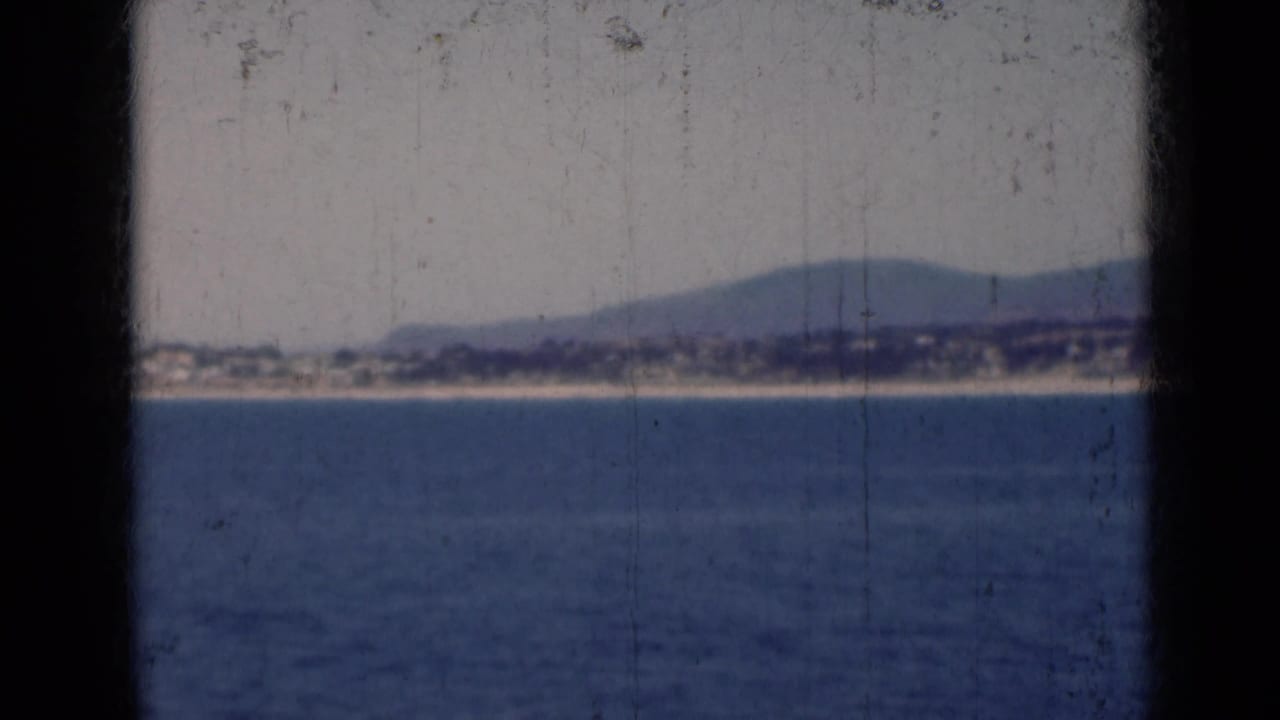 Calm blue sea with a distant shoreline and low hills behind dark 8mm frame borders.