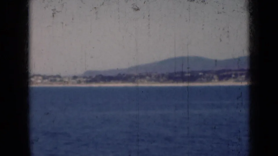1947 India 8mm frame showing calm water, a pale coast, low distant buildings, hazy hills, and visible film wear.
