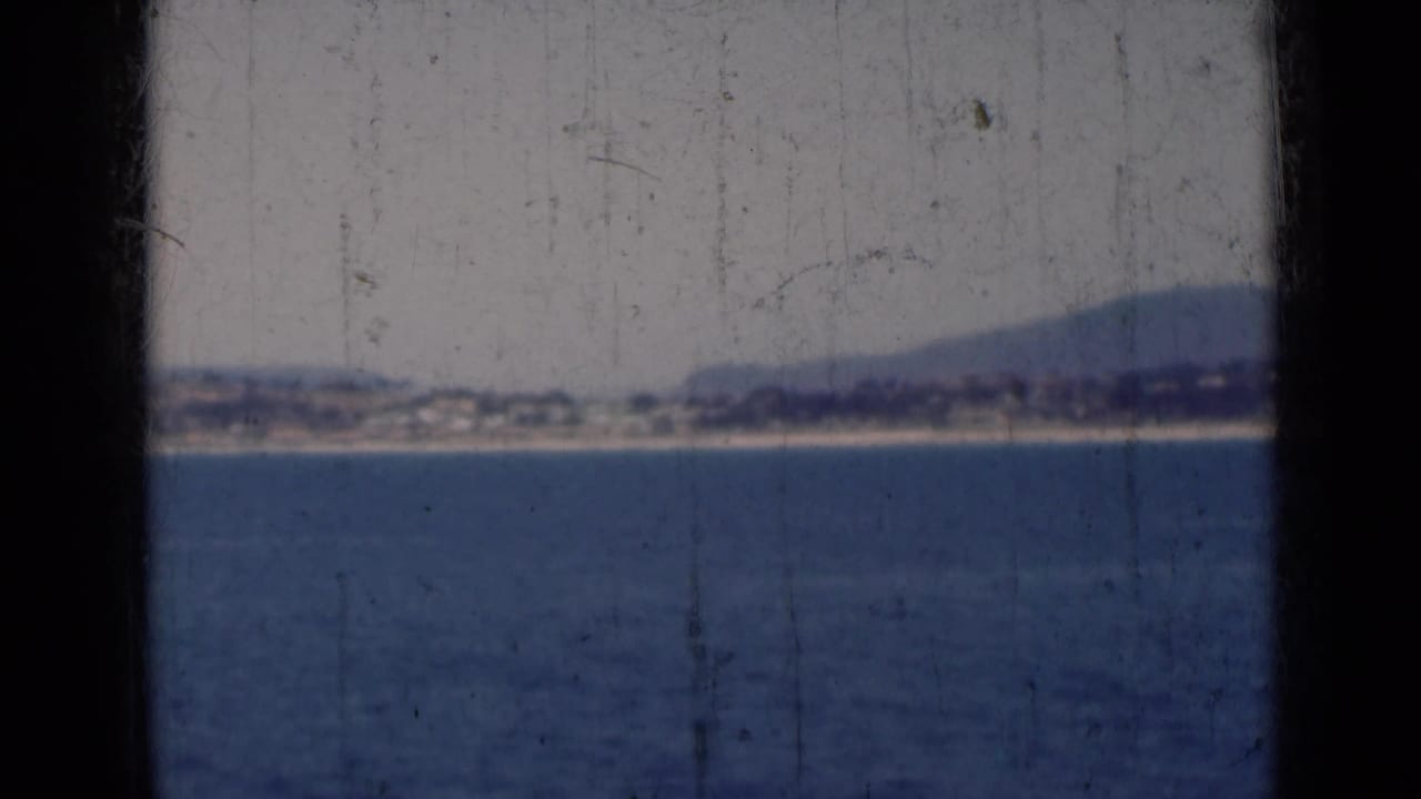 Distant coast across blue water, with low hills and visible scratches on the film surface.