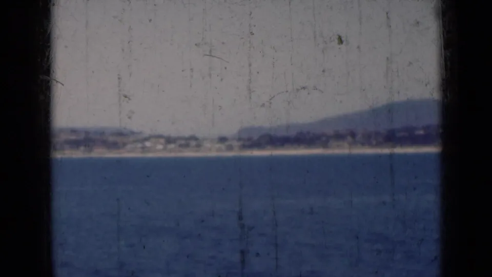 A grainy 8mm view across the sea toward a low Indian shoreline, with hills in the background and heavy film markings.