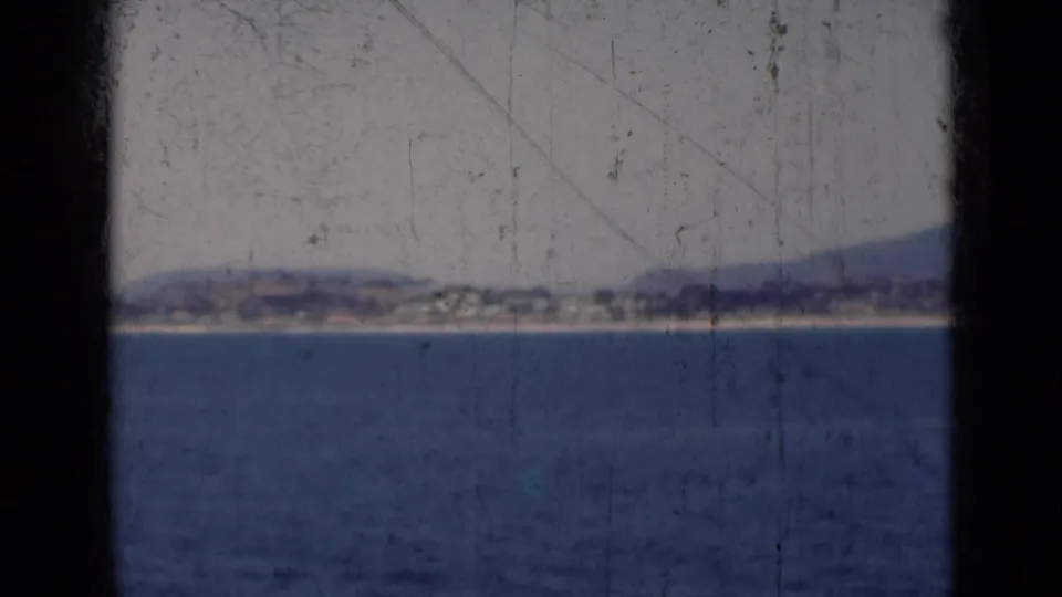 1947 India home-movie frame of open water leading to a distant coast, softened by haze, grain, dust, and scratches.