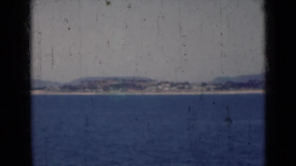 A soft 8mm coastal view in India from 1947, showing calm sea, a distant shoreline, low hills, and aged film texture.