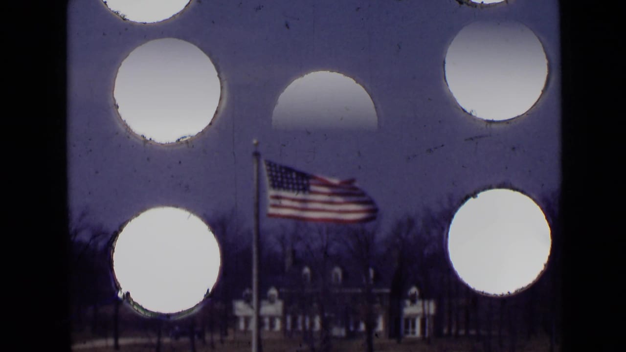 American flag on a pole seen through circular foreground openings with trees and a distant building behind it.