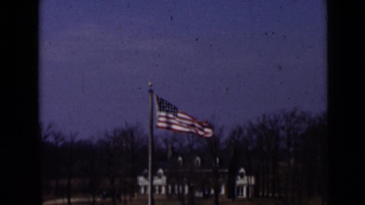 1947 New York 8mm frame of an American flag fluttering on a flagpole, with bare trees and a light-colored building visible beyond the lawn.