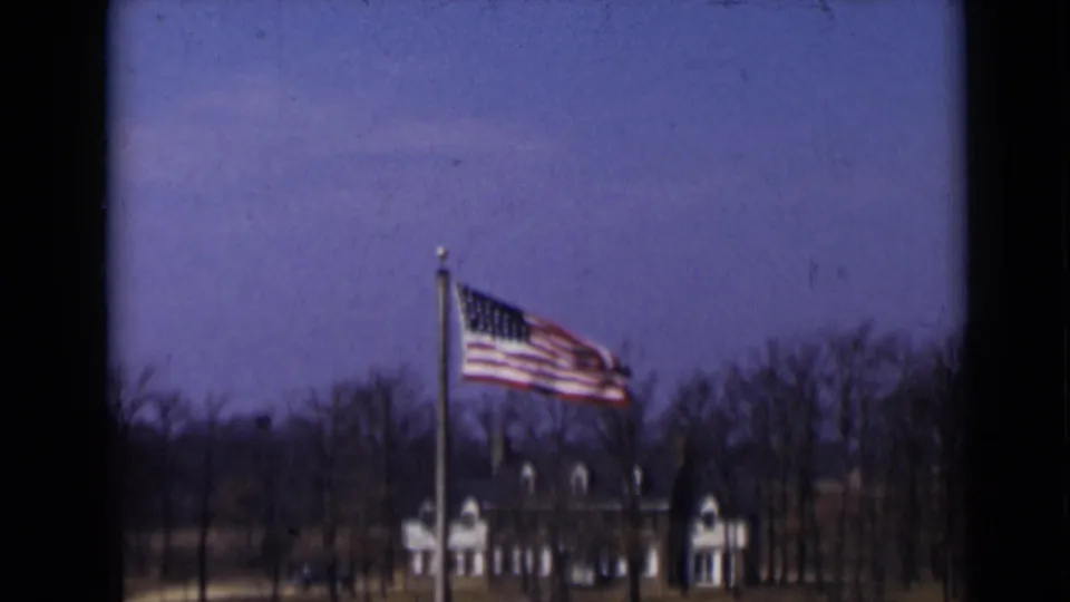 1947 New York 8mm frame showing the American flag in wind, a distant building with dormer-like roof details, bare trees, and dark film edges.