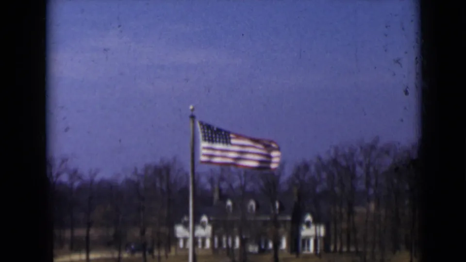 1947 New York 8mm frame with an American flag waving steadily, bare trees across the background, and a distant light-colored building below a blue sky.