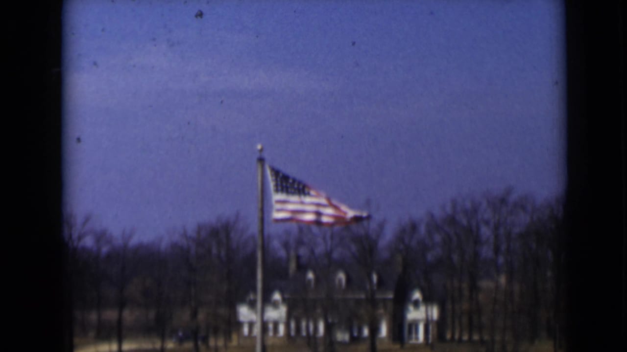 A U.S. flag waves in wind in 1947 New York, with bare trees and a large building visible beyond the pole.