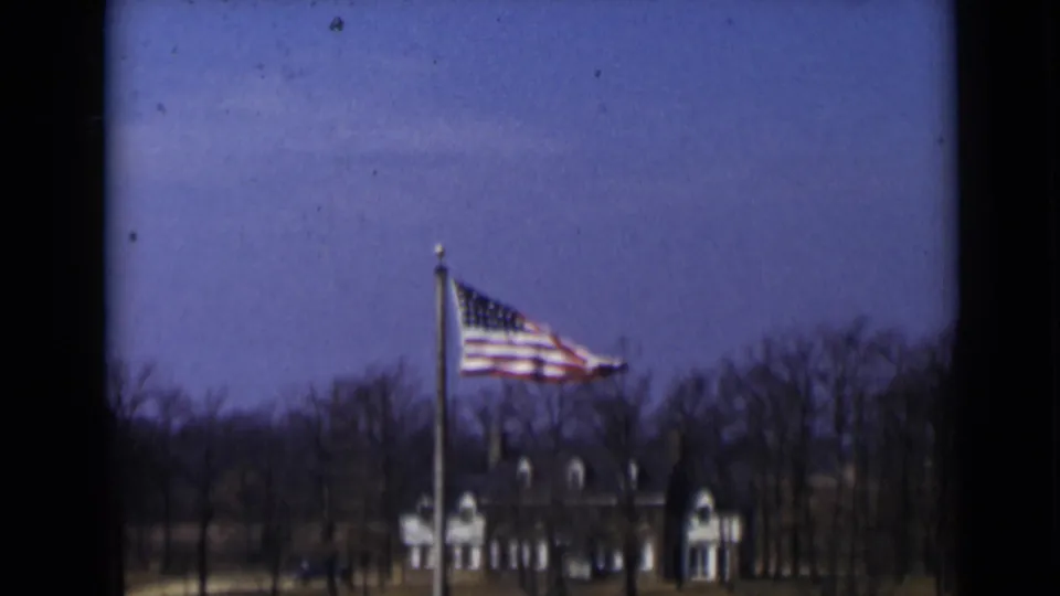 A U.S. flag waves in wind in 1947 New York, with bare trees and a large building visible beyond the pole.