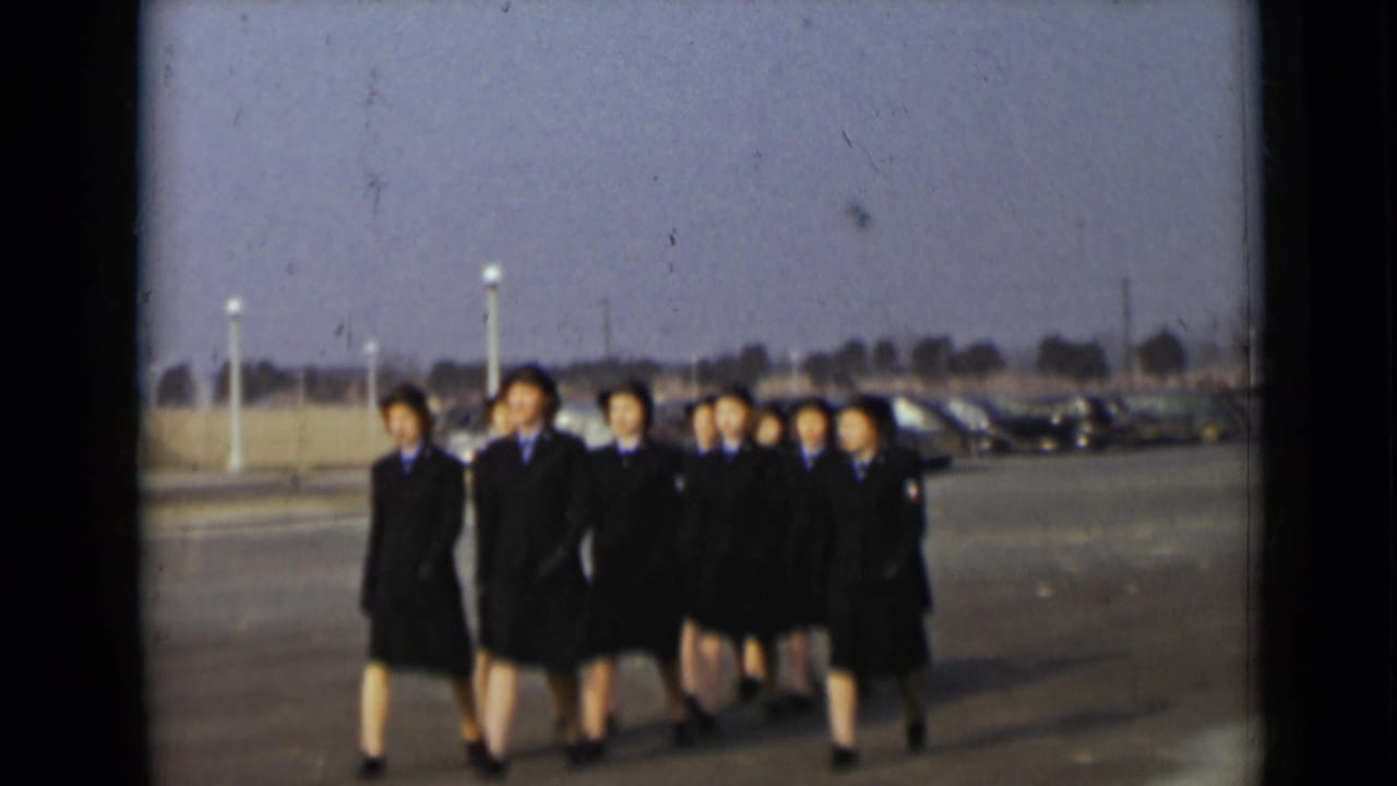 Women in dark uniforms and hats march toward the camera across a paved lot.