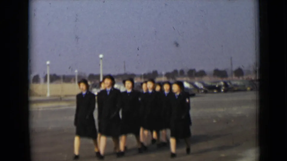A group of women in dark uniform coats and hats marches in formation across a paved lot in New York, 1947.