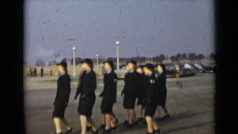 Women in dark service-style uniforms and hats move in formation across a paved New York lot in 1947.