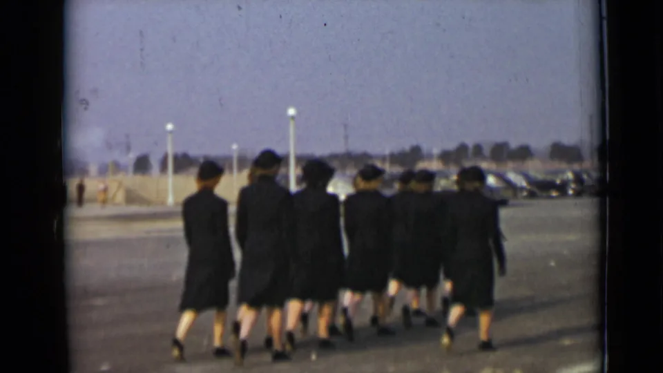 A formation of women in dark uniforms marches away across an open paved lot in 1947 New York.