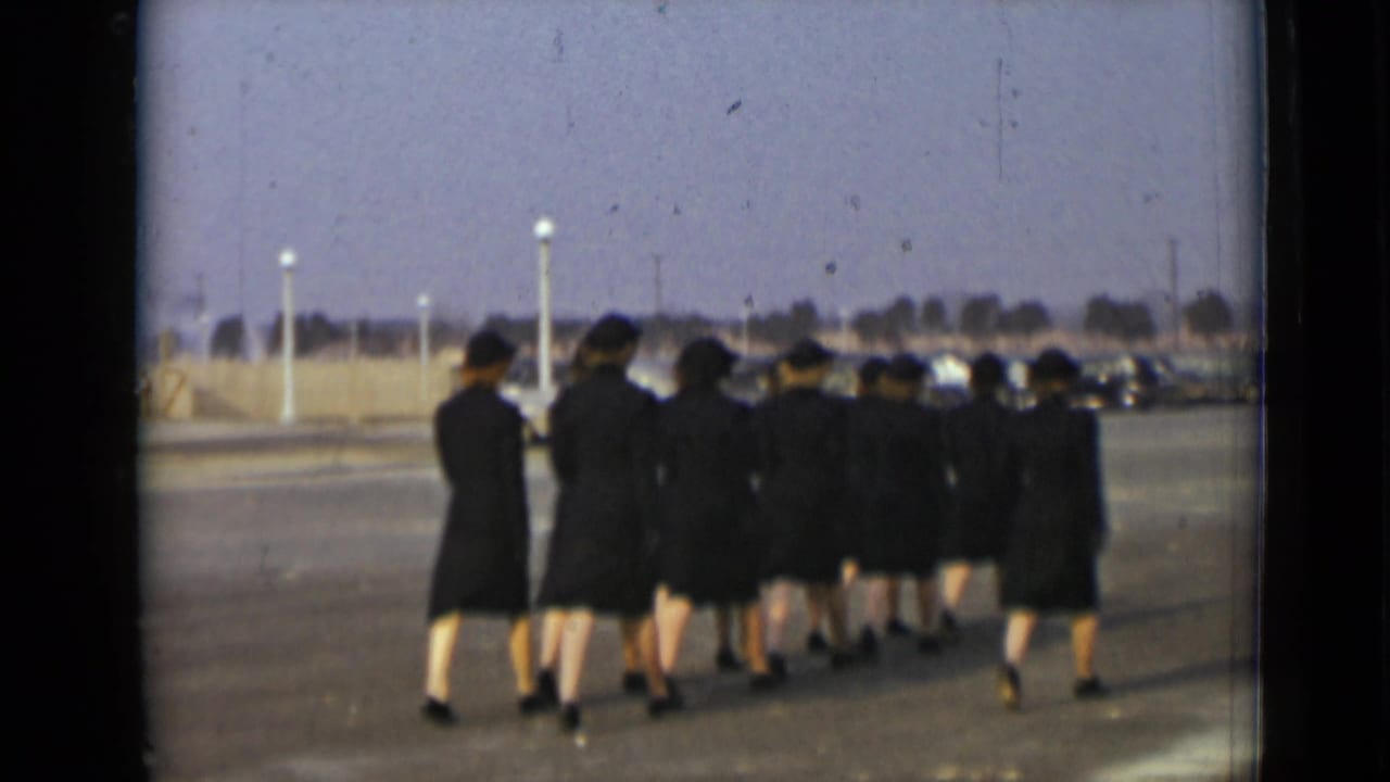 Women in dark uniforms walk away across a paved lot with cars in the background.