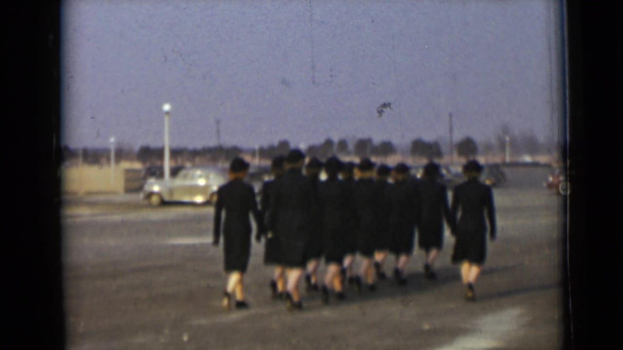 Uniformed women in dark hats and coats march away in formation across a paved lot, with a light-colored car visible behind them.