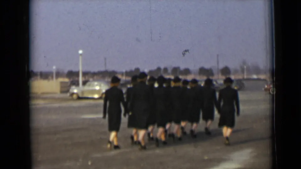 Uniformed women in dark hats and coats march away in formation across a paved lot, with a light-colored car visible behind them.