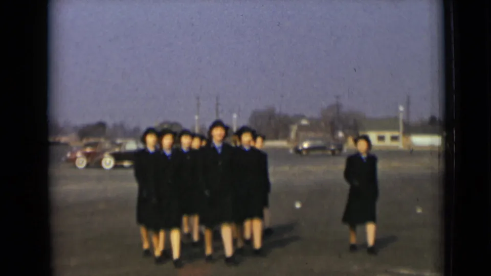 Women in matching dark uniforms and hats approach the camera across a paved New York lot, with older cars and low buildings in the background.