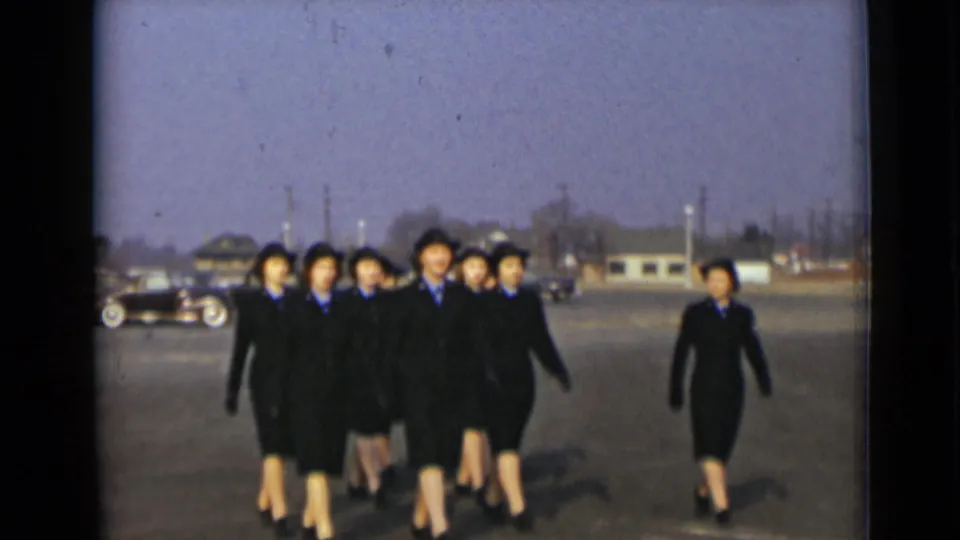 The formation of uniformed women advances toward the camera, while a separate uniformed woman walks along the right side of the paved lot.