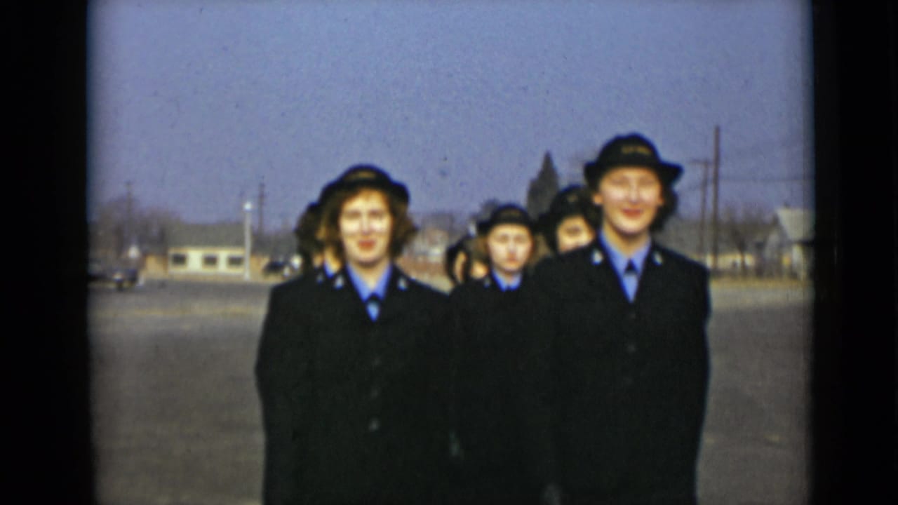 Uniformed young women march toward the camera on a paved road.