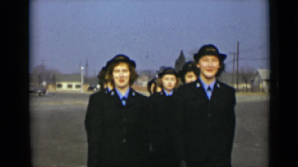 In 1947 New York footage, young women in dark uniforms and hats march toward the camera on a suburban road.