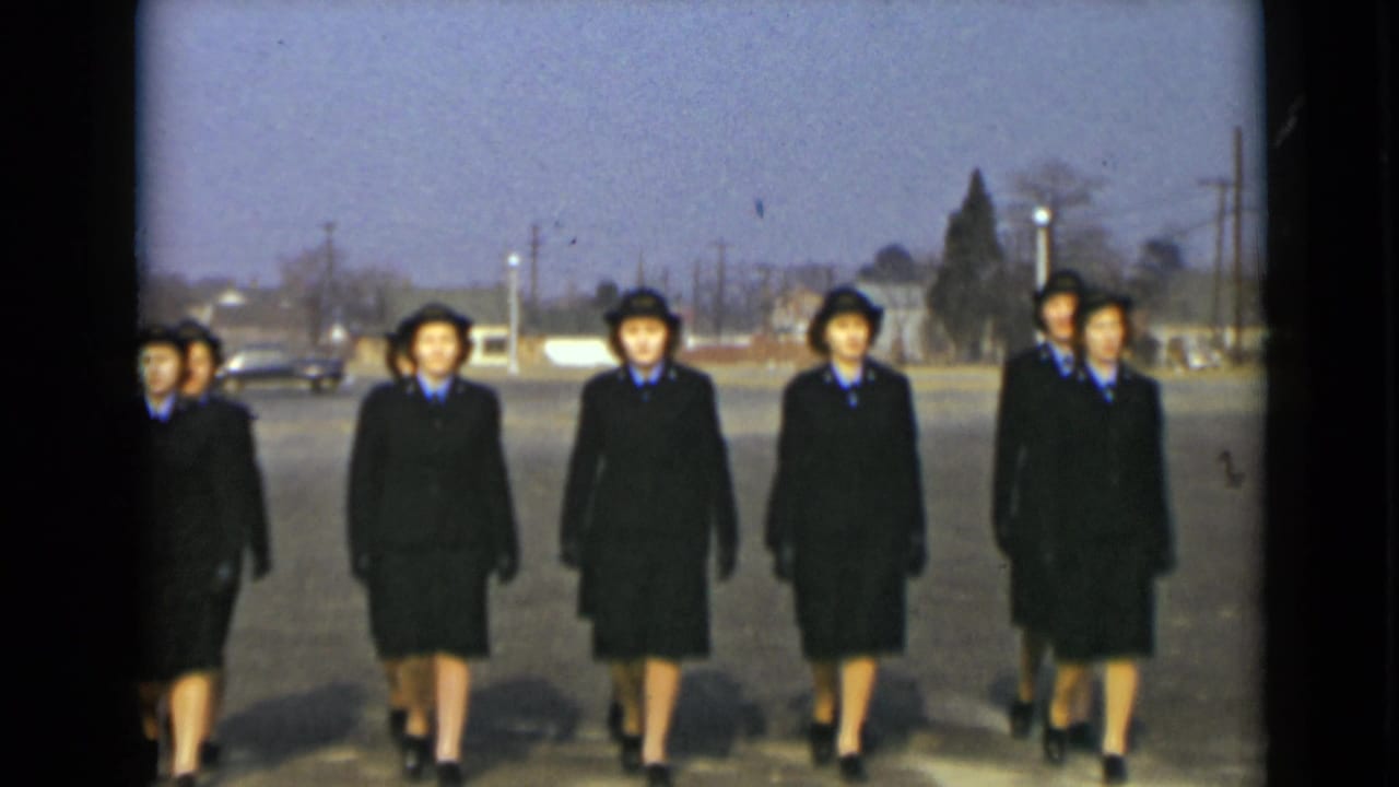 Women in dark uniforms and hats march toward the camera across a paved lot.