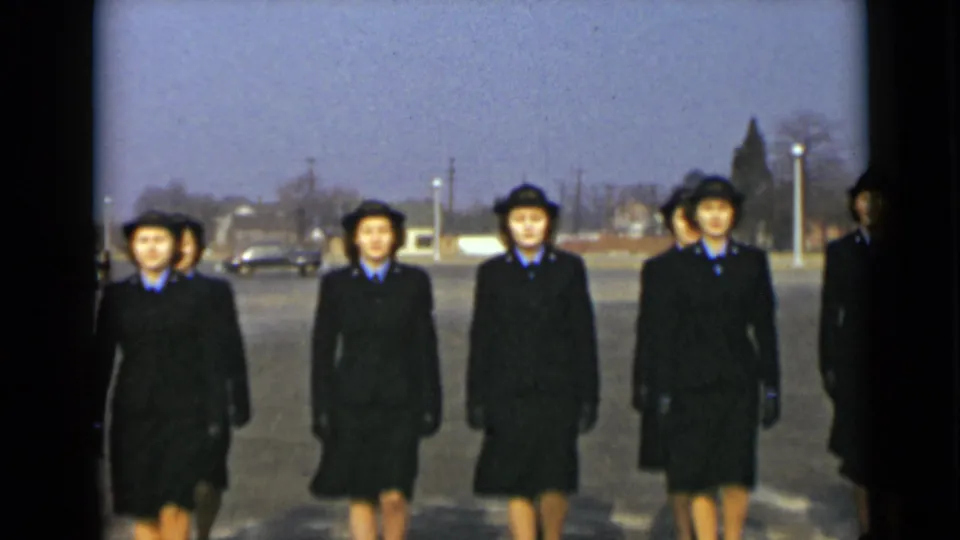 Women in matching dark uniforms and hats approach the camera in a New York 1947 outdoor drill scene.