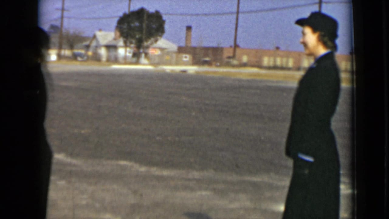 Woman in a dark uniform and hat stands in profile beside a paved lot.