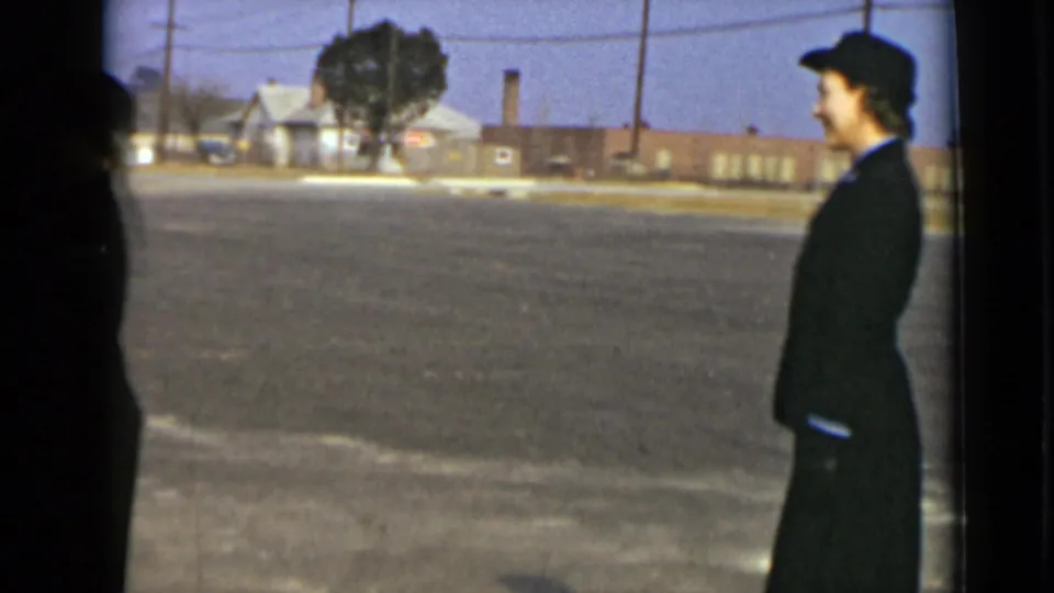 1947 New York 8mm frame showing a woman in a dark skirted uniform and hat in profile near an open paved lot.