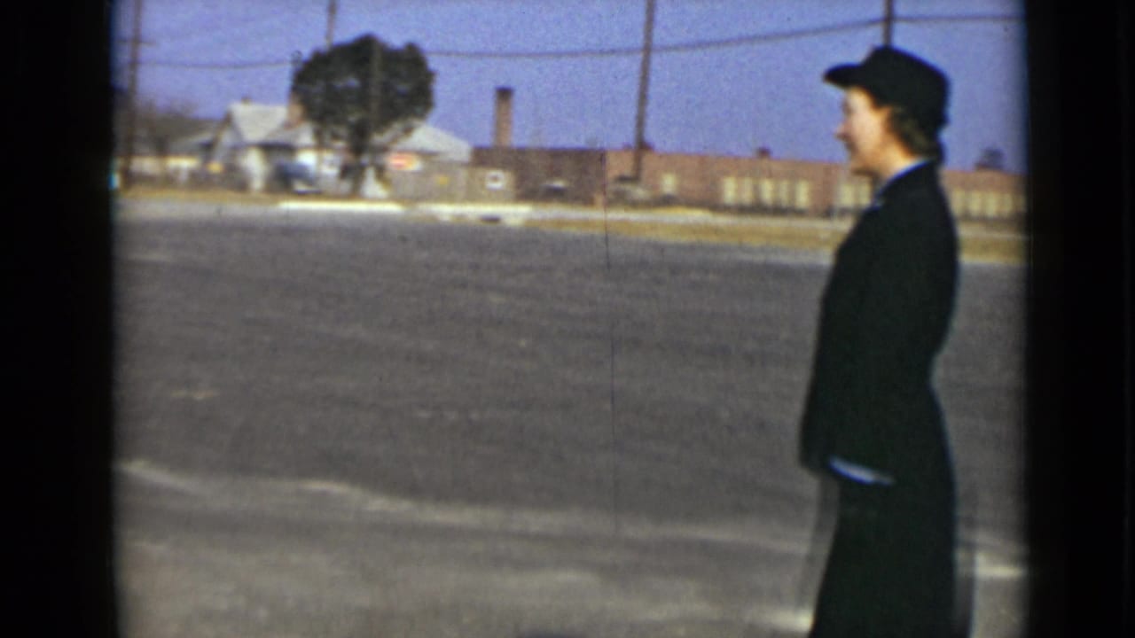 A woman in a dark uniform and hat is seen in profile against a paved lot and low New York buildings in 1947 footage.