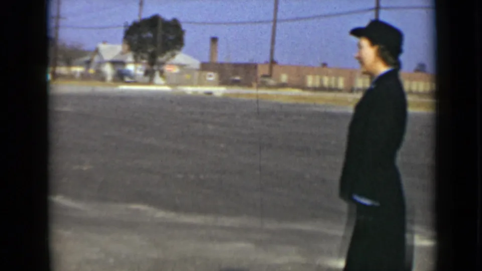 A woman in a dark uniform and hat is seen in profile against a paved lot and low New York buildings in 1947 footage.