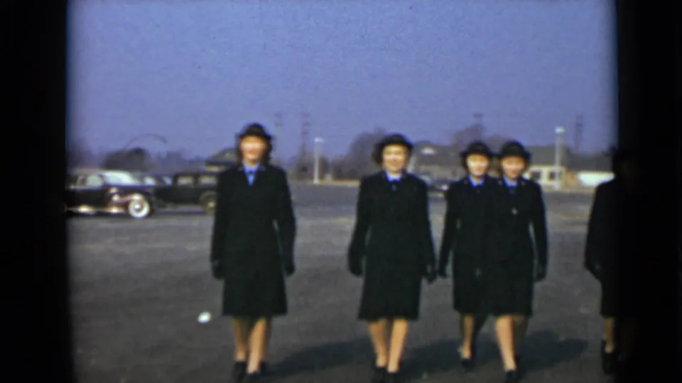 Uniformed women walk in formation across a paved New York lot, with a classic car and low buildings behind them.