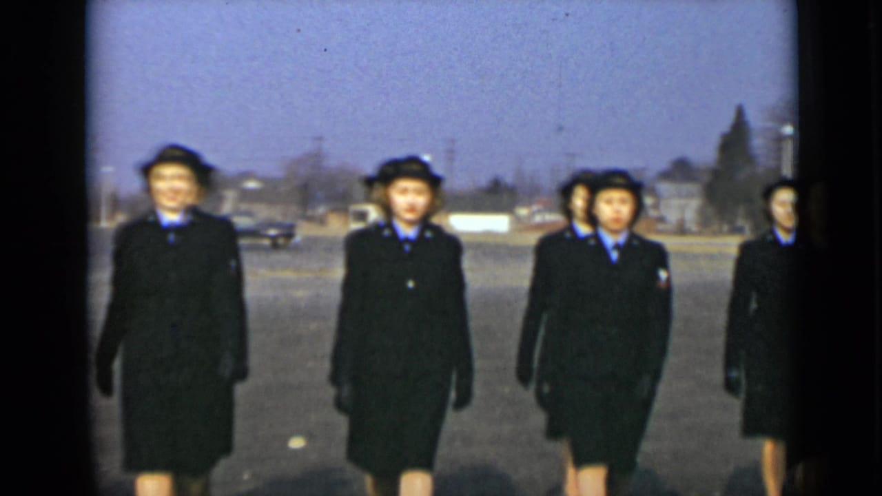 Uniformed women in dark coats and hats march across a paved open area.