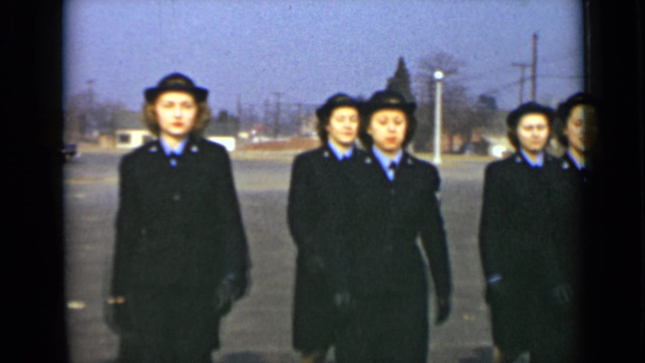 Uniformed women in matching dark coats and hats approach the camera in formation, with utility poles and low buildings behind them.