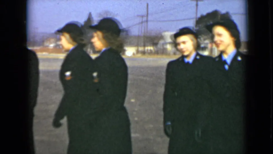 Uniformed women in dark coats and hats move past the camera, some looking sideways as the formation shifts across the paved lot.