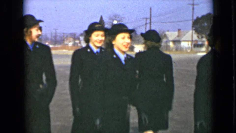 Women in matching dark uniforms stand or turn together outdoors, several smiling as the home-movie camera records them.