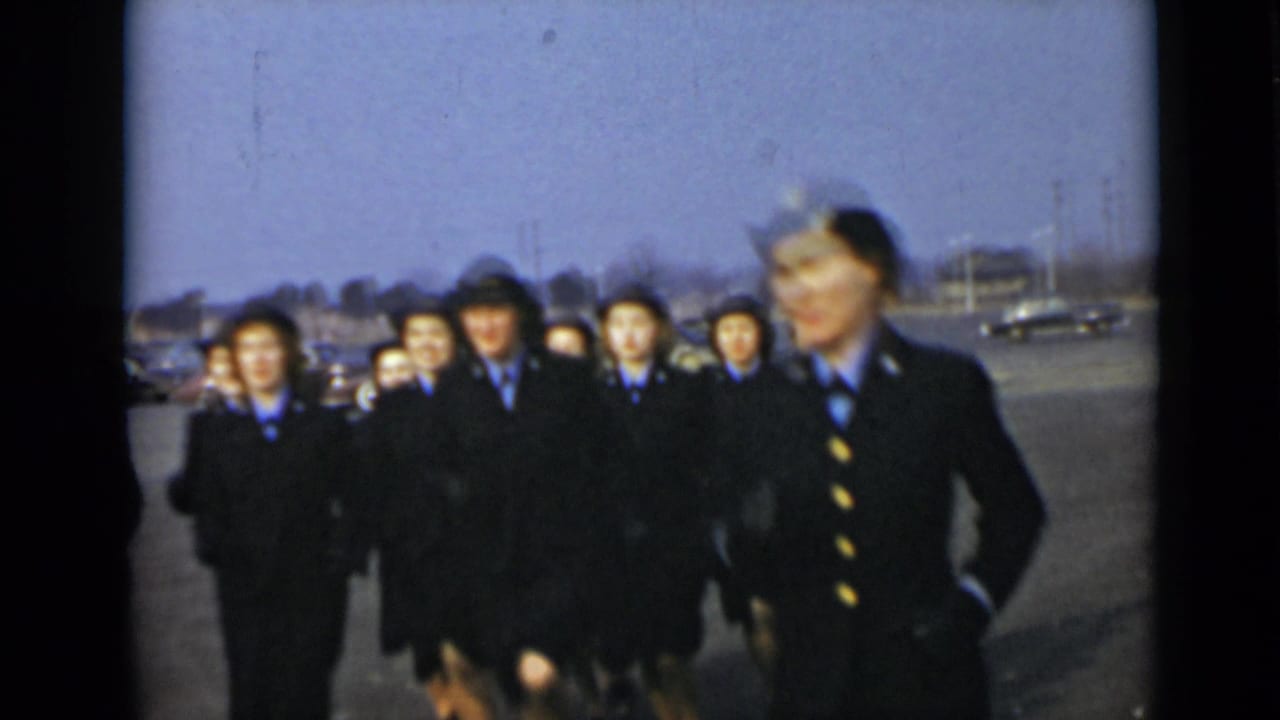 Uniformed women walk in formation across an open lot with parked cars behind them.