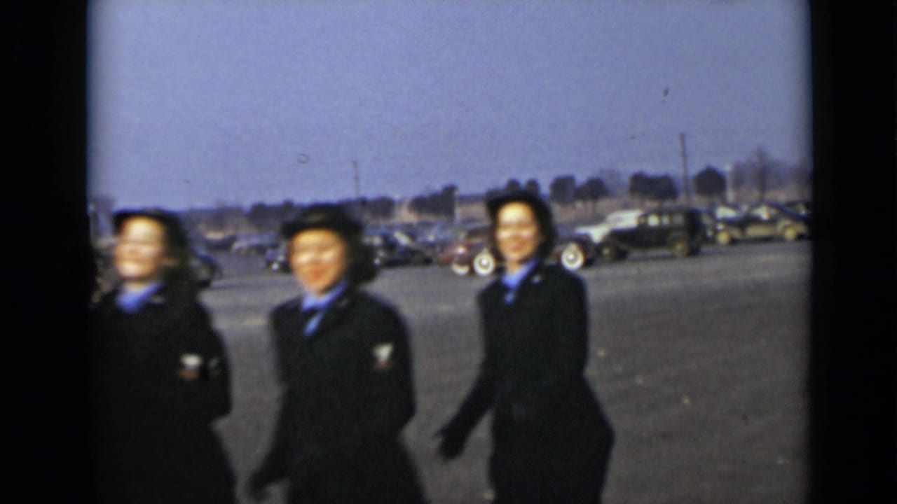 Three women in dark blue uniforms and hats pass through the frame in New York, with mid-century cars behind them.