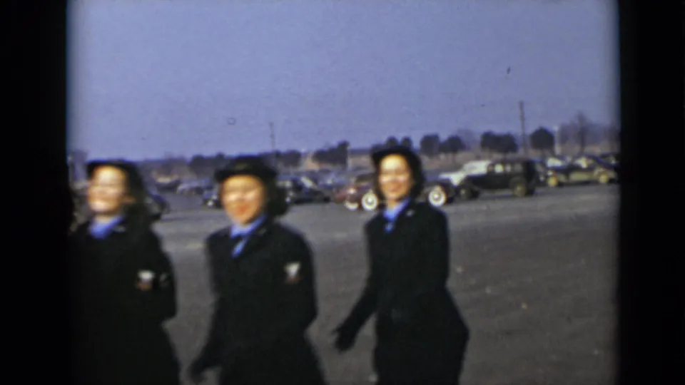 Three women in dark blue uniforms and hats pass through the frame in New York, with mid-century cars behind them.
