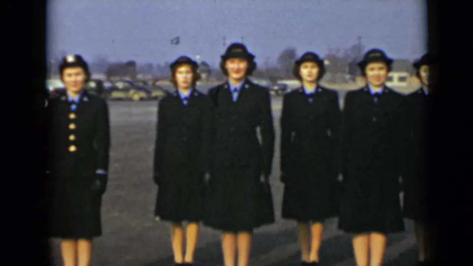 Uniformed women stand in formation for the camera, with coats, hats, blue collars, and parked cars visible behind them.