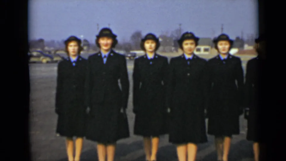 Five women in matching dark uniforms and hats pose in a neat outdoor line in the 1947 New York footage.
