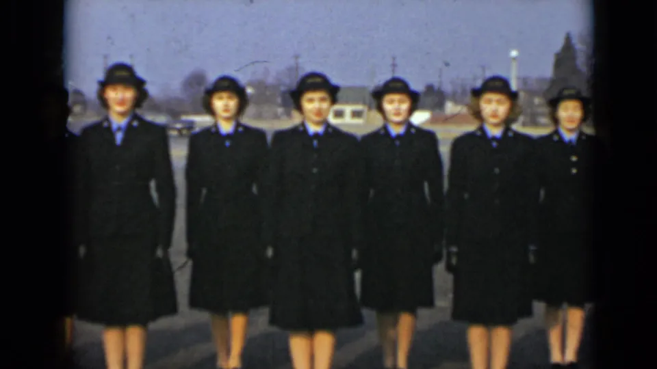 Six women in matching dark uniforms face the camera in an outdoor New York setting, with parked vehicles and utility poles in the background.