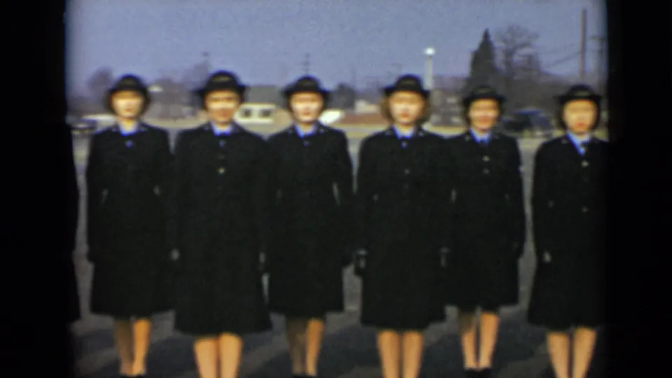 A steady 1947 home-movie frame shows six women in dark uniform coats and hats standing shoulder to shoulder outside in New York.