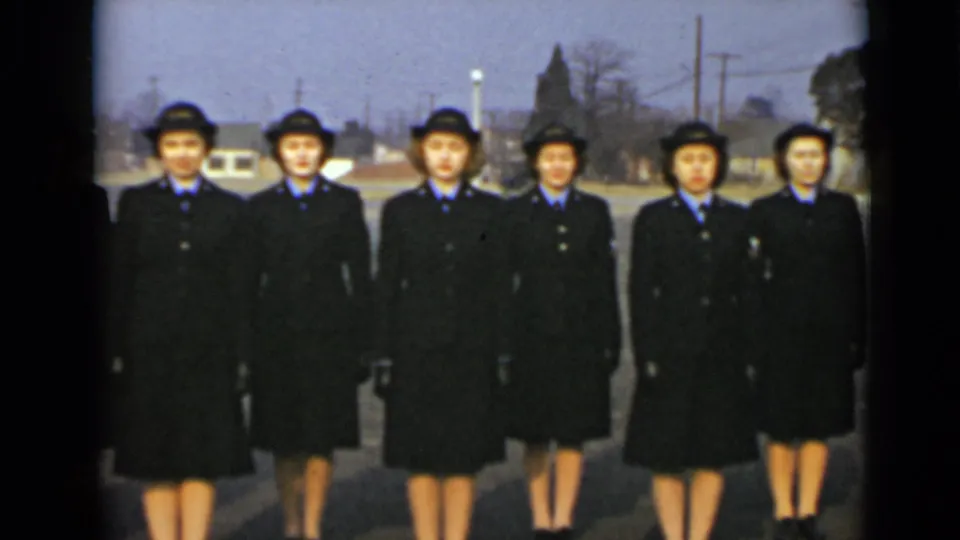 Six women in military-style dress stand in a formal line outdoors, framed by late-1940s New York roadside details.