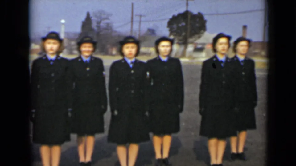 Women in matching blue military-style uniforms stand together outdoors in New York, with utility poles and buildings behind them.